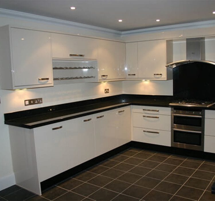 L-shape modular kitchen 2 Elegant l-shaped modular kitchen with glossy white cabinetry and black countertops. The kitchen features under-cabinet lighting, a built-in oven, a chimney with a black backsplash, and modern handles for a sleek, contemporary look. The dark tiled flooring adds contrast and sophistication to the space.