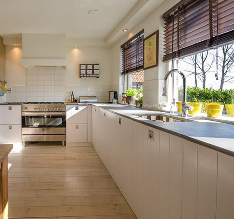 L-shape modular kitchen 4 Bright and airy l-shaped modular kitchen with white wooden cabinets and a black countertop. The space features a large stainless steel gas stove, a farmhouse-style sink, and wide windows with wooden blinds, allowing ample natural light. The warm wooden flooring and yellow potted plants enhance the cozy and inviting ambiance.