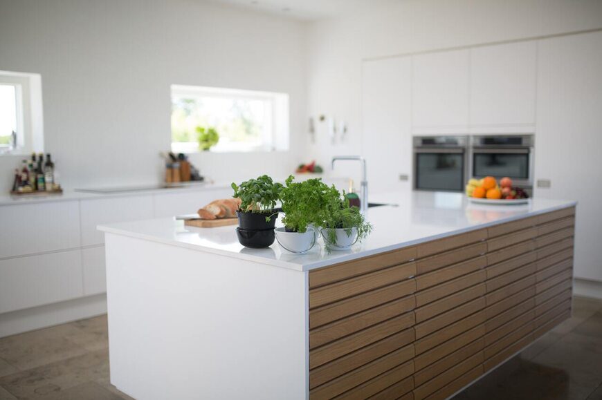 Modern island modular kitchen by alea modular kitchen featuring a sleek white countertop, built-in sink, and wooden cabinetry for a stylish and functional cooking space. The kitchen is well-lit with natural light, complemented by fresh herbs and fruits for a contemporary aesthetic.